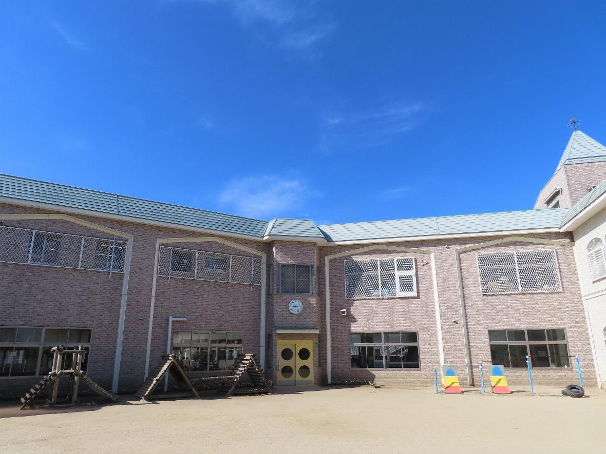 園庭から見た園舎正面。中央に丸い窓のついた黄色い扉と壁時計があり、左右に保育室の大きな窓が並んでいる。